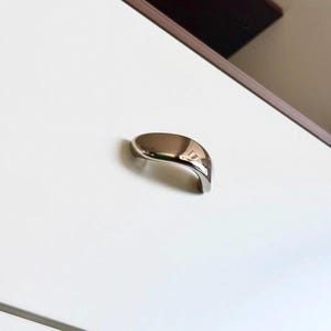 May include: A polished silver-toned metal drawer pull with a smooth, curved, arching design. The hardware features a reflective, metallic finish and is mounted onto a flat, white surface, showcasing a clean, simple, and functional aesthetic.