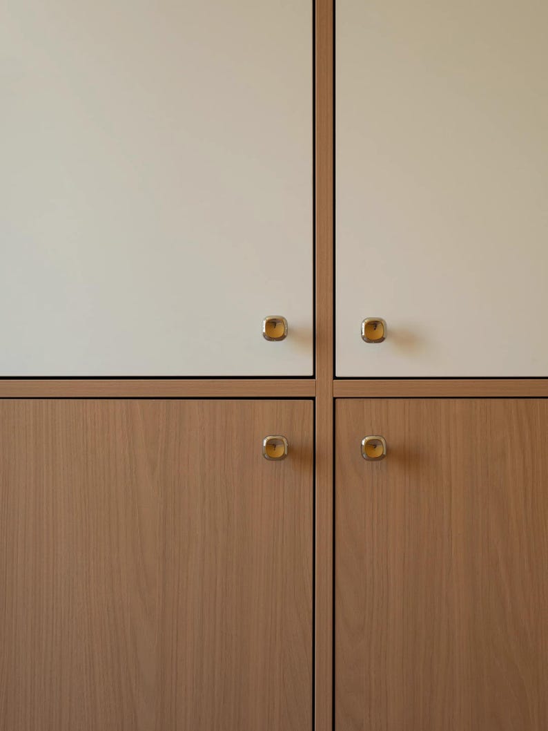 Puede incluir: Un armario de almacenamiento con cuatro puertas dispuestas en cuadr&iacute;cula. Las dos puertas superiores son de color blanco roto mate, mientras que las dos inferiores muestran un acabado de veta de madera clara natural. Cada puerta tiene un pomo met&aacute;lico cuadrado.