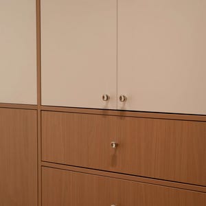 Puede incluir: Una unidad de almacenamiento con dos puertas de armario superiores en acabado blanco roto mate y tres cajones inferiores con textura de veta de madera c&aacute;lida. Cada puerta y caj&oacute;n tiene un peque&ntilde;o pomo redondo de metal plateado.