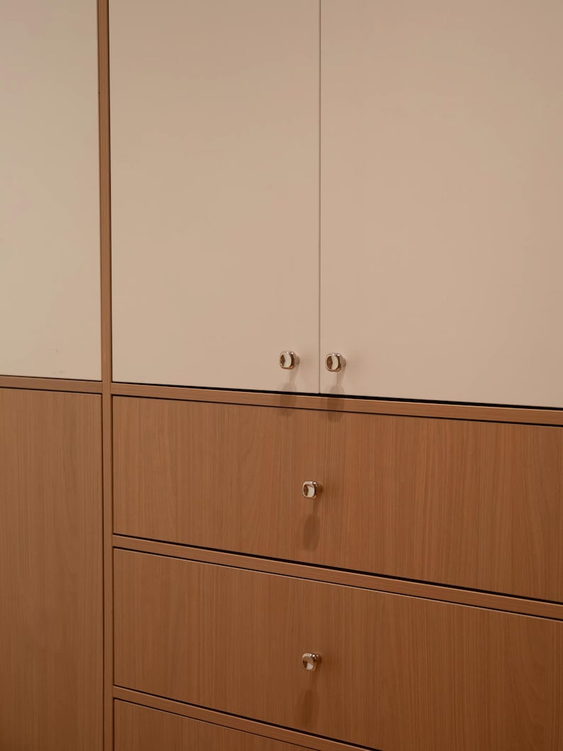 Puede incluir: Una unidad de almacenamiento con dos puertas de armario superiores en acabado blanco roto mate y tres cajones inferiores con textura de veta de madera c&aacute;lida. Cada puerta y caj&oacute;n tiene un peque&ntilde;o pomo redondo de metal plateado.