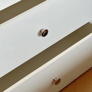 Puede incluir: Una c&oacute;moda blanca con dos cajones abiertos de superficies lisas y mate. Cada caj&oacute;n tiene un pomo circular. El pomo superior presenta un centro marr&oacute;n oscuro pulido, mientras que el pomo inferior es de metal plateado con acabado reflectante.