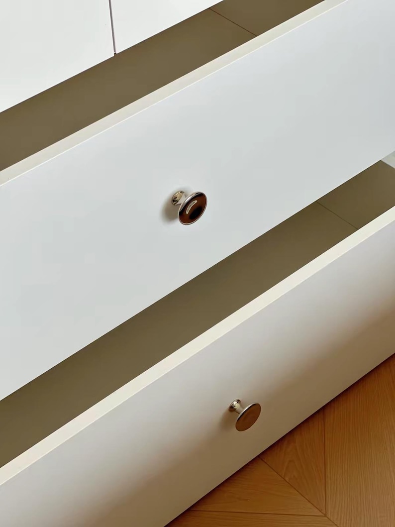 Puede incluir: Una c&oacute;moda blanca con dos cajones abiertos de superficies lisas y mate. Cada caj&oacute;n tiene un pomo circular. El pomo superior presenta un centro marr&oacute;n oscuro pulido, mientras que el pomo inferior es de metal plateado con acabado reflectante.