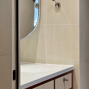 Puede incluir: Un mueble de ba&ntilde;o con encimera blanca y armarios de color blanco roto con bordes de madera oscura. Incluye pomos plateados, un espejo ovalado alto y dos ganchos de pared con esferas de madera sobre azulejos de color crema.