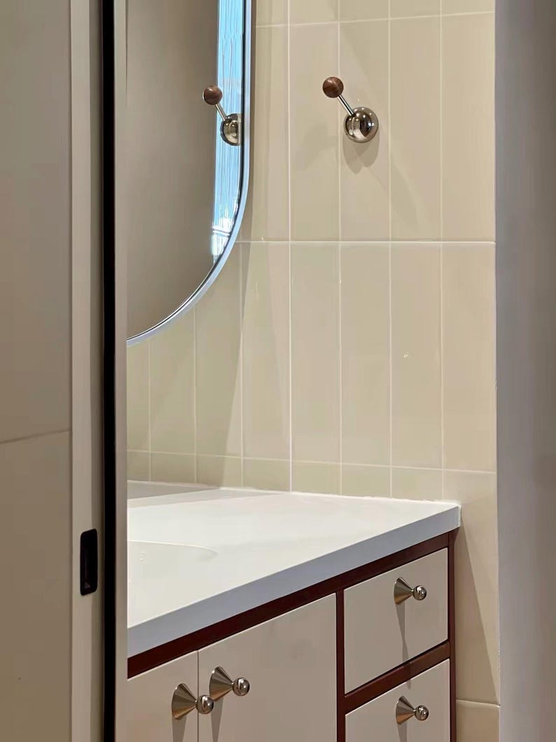 Puede incluir: Un mueble de ba&ntilde;o con encimera blanca y armarios de color blanco roto con bordes de madera oscura. Incluye pomos plateados, un espejo ovalado alto y dos ganchos de pared con esferas de madera sobre azulejos de color crema.