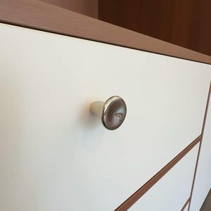 Puede incluir: Un pomo de mueble circular de cromo pulido con un centro reflectante ligeramente c&oacute;ncavo, montado sobre un frente de caj&oacute;n blanco mate plano que presenta un borde fino de madera clara a lo largo del marco del mueble.