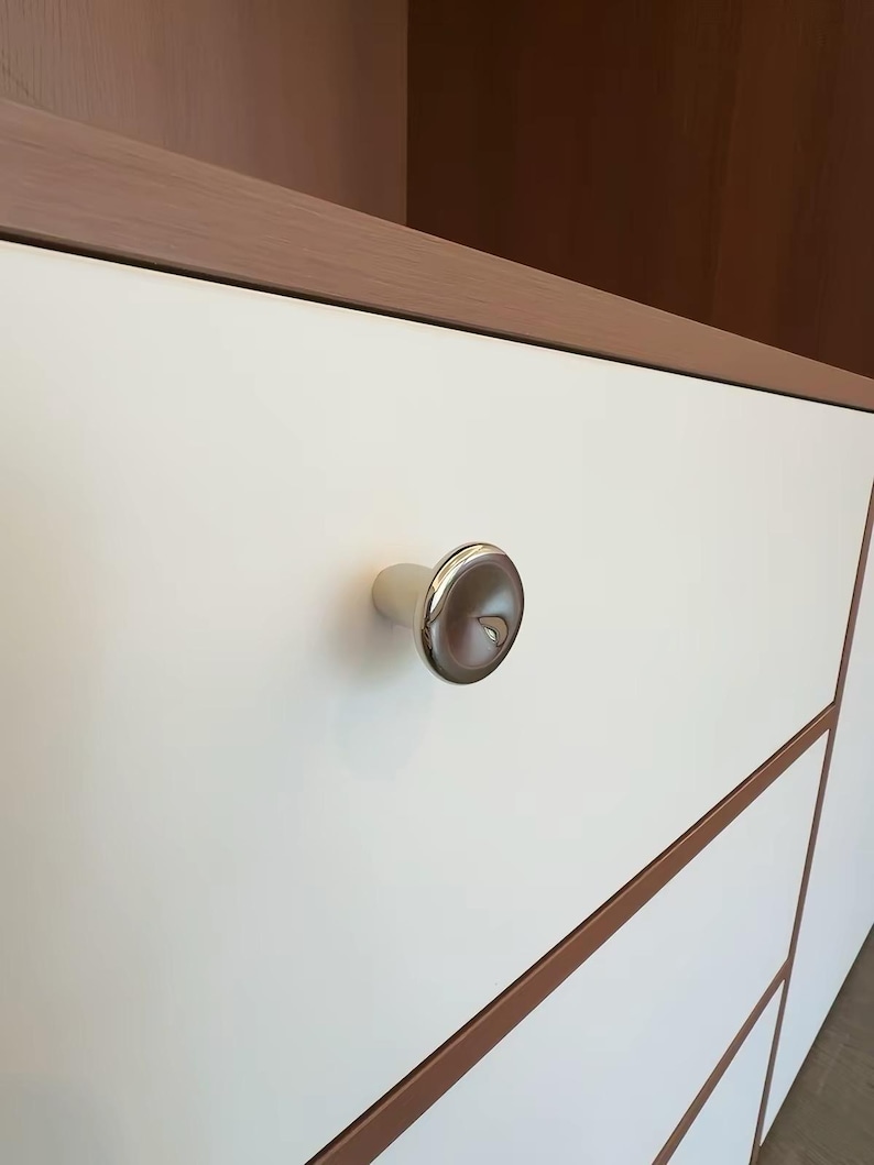 Puede incluir: Un pomo de mueble circular de cromo pulido con un centro reflectante ligeramente c&oacute;ncavo, montado sobre un frente de caj&oacute;n blanco mate plano que presenta un borde fino de madera clara a lo largo del marco del mueble.