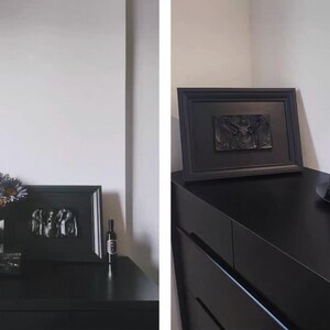 K&ouml;nnte beinhalten: Eine schwarze Kommode mit gerahmter Kunst und dekorativen Gegenst&auml;nden. Ein Rahmen zeigt eine Reliefskulptur. Eine Vase mit Blumen und eine Flasche befinden sich ebenfalls auf der Kommode. Eine schwarze Skulptur steht auf einem schwarzen Teller.