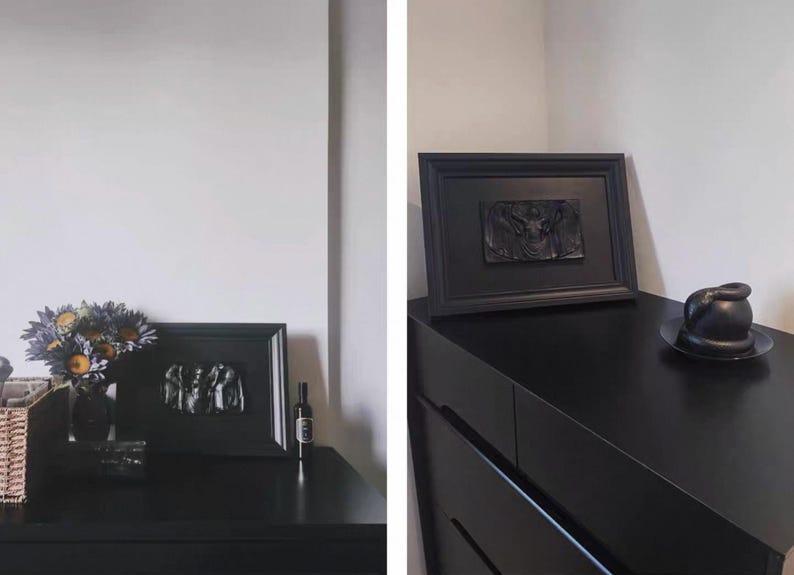 K&ouml;nnte beinhalten: Eine schwarze Kommode mit gerahmter Kunst und dekorativen Gegenst&auml;nden. Ein Rahmen zeigt eine Reliefskulptur. Eine Vase mit Blumen und eine Flasche befinden sich ebenfalls auf der Kommode. Eine schwarze Skulptur steht auf einem schwarzen Teller.