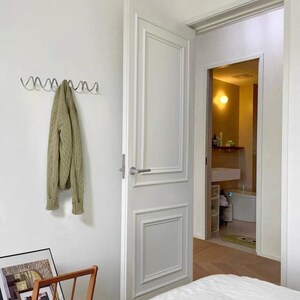 May include: A light-filled bedroom with a white door and a glimpse into a bathroom. A green sweater hangs on a silver wall hook. A wooden magazine rack holds framed pictures, and a bed with white linens is in the foreground.