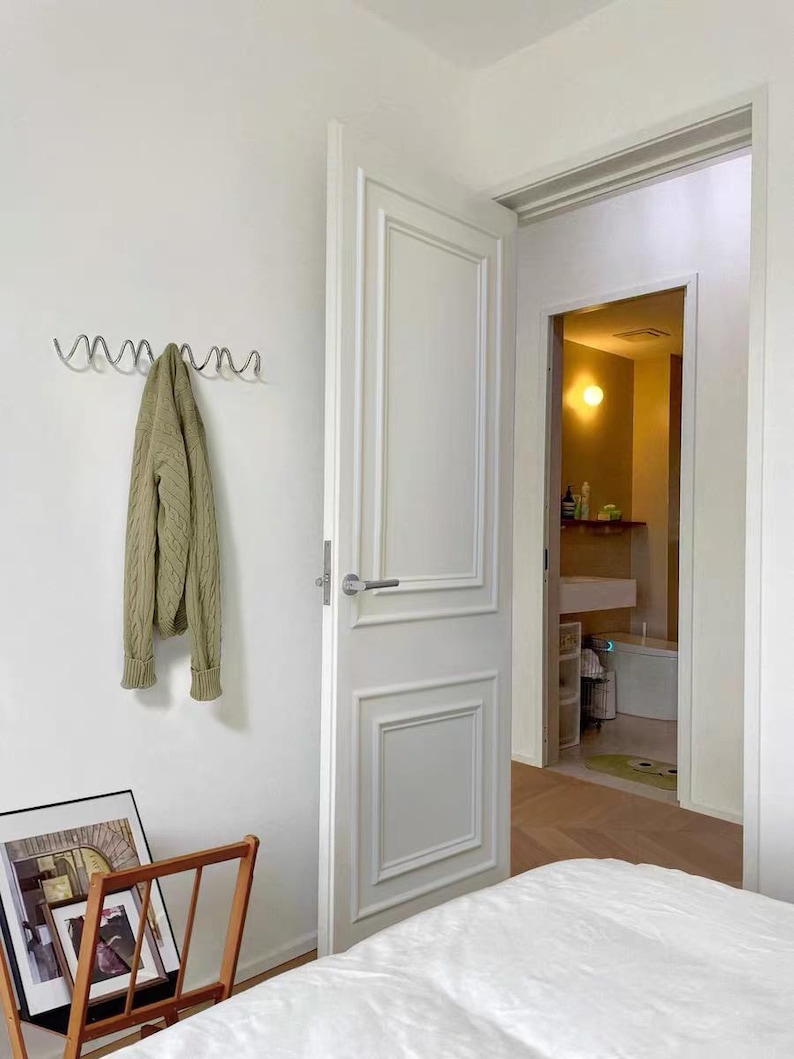 May include: A light-filled bedroom with a white door and a glimpse into a bathroom. A green sweater hangs on a silver wall hook. A wooden magazine rack holds framed pictures, and a bed with white linens is in the foreground.