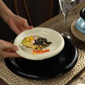 May include: A white ceramic plate holding a prepared dish with avocado, corn, and dark-colored food. The plate sits atop a black plate, with a glass of water and a blue plate in the background. The food is arranged on a wooden table.