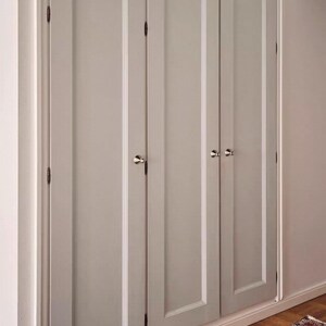 Puede incluir: Un conjunto de tres puertas de armario de color gris claro con paneles empotrados verticales y peque&ntilde;os pomos redondos de metal plateado. Las puertas est&aacute;n montadas con bisagras de metal oscuro y enmarcadas por un borde blanco.