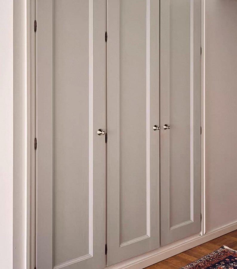 Puede incluir: Un conjunto de tres puertas de armario de color gris claro con paneles empotrados verticales y peque&ntilde;os pomos redondos de metal plateado. Las puertas est&aacute;n montadas con bisagras de metal oscuro y enmarcadas por un borde blanco.