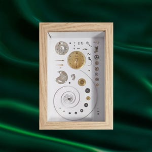 Peut inclure: Une œuvre encadrée présentant des composants de montre sur fond blanc. On y trouve un cadran doré, des engrenages, des vis et un ressort en spirale. Le cadre est en bois clair.