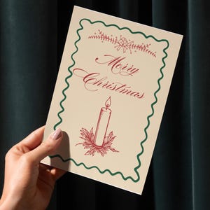 Może przedstawiać: Kremowa kartka świąteczna z zieloną falistą obwódką i czerwonym napisem "Merry Christmas". Karta zawiera ilustrację świecy z czerwonym płomieniem i liśćmi ostrokrzewu. Karta jest trzymana na tle ciemnozielonej zasłony.