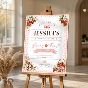 Puede incluir: Un cartel de cumpleaños rectangular sobre un caballete de madera con un borde de cuadros vichy rosa y encaje. El texto dice: Welcome to Jessica's One Sweet Year Berry First Birthday September 17, 2026. Decorado con ilustraciones de fresas y lazos.