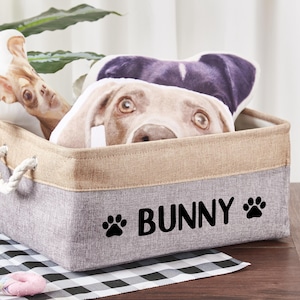 May include: A rectangular storage bin with a gray bottom and tan top, featuring the word "BUNNY" and paw prints. The bin contains dog-themed plush toys, including a bone and a dog's face.