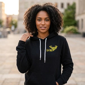 Può includere: Felpa nera con coulisse bianche e tasca frontale. La felpa presenta la scritta "LA-TAZ" in giallo con un disegno a vortice giallo. La persona indossa la felpa all'aperto.