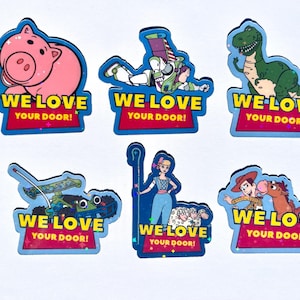 May include: A set of six colorful stickers featuring animated characters including a pink pig, a space ranger, a green dinosaur, a race car, a shepherdess with sheep, and a cowboy with a horse. Each sticker displays the text WE LOVE YOUR DOOR!