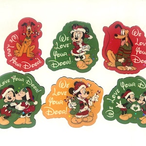 May include: Six die-cut stickers featuring Disney characters in holiday attire. Each sticker displays the text "We Love Your Door!" and depicts Pluto, Minnie Mouse, or Mickey Mouse in festive scenes with candy canes, Christmas lights, and Santa hats.