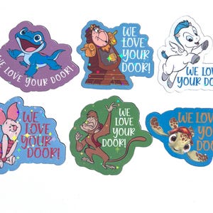 May include: A set of six colorful stickers featuring animated characters including a blue lizard, a clock, a white winged horse, a pink pig, a brown monkey, and a sea turtle. Each sticker includes the text WE LOVE YOUR DOOR in blue or white.