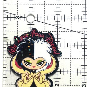 Puede incluir: Un dije de estilo caricatura con un personaje de cabello negro y blanco, ojos verdes y labio rojo. El dije tiene un atuendo amarillo y negro y el texto "We love your door!" est&aacute; escrito en la parte superior.