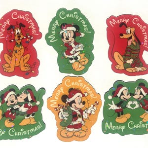 May include: A set of six die-cut stickers featuring Disney characters in holiday attire. Each sticker displays the text Merry Christmas and depicts Pluto, Minnie Mouse, or Mickey Mouse with candy canes, string lights, or Santa hats on red, green, or orange backgrounds.