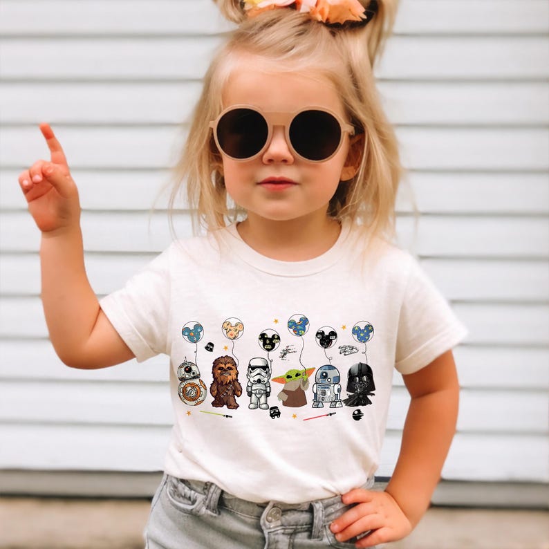 K&ouml;nnte beinhalten: Cremefarbenes Kurzarm-T-Shirt mit einem farbenfrohen Aufdruck von Star Wars-Charakteren und Luftballons. Das Design umfasst BB-8, Chewbacca, einen Sturmtruppler, Grogu, R2-D2 und Darth Vader. Die Ballons haben Mickey Mouse Ohren.