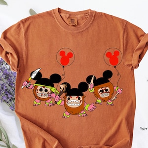 May include: Burnt orange t-shirt featuring a cartoon design of three characters with Mickey Mouse ears, holding weapons and balloons. The characters have brown, pink, and green accents. The balloons are red.