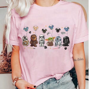 K&ouml;nnte beinhalten: Hellrosa T-Shirt mit Star Wars-Figuren, darunter BB-8, Chewbacca, ein Sturmtruppler, Grogu, R2-D2 und Darth Vader. Jede Figur wird mit einem Mickey Mouse-Ballon dargestellt. Das Design beinhaltet Sterne und Lichtschwerter.