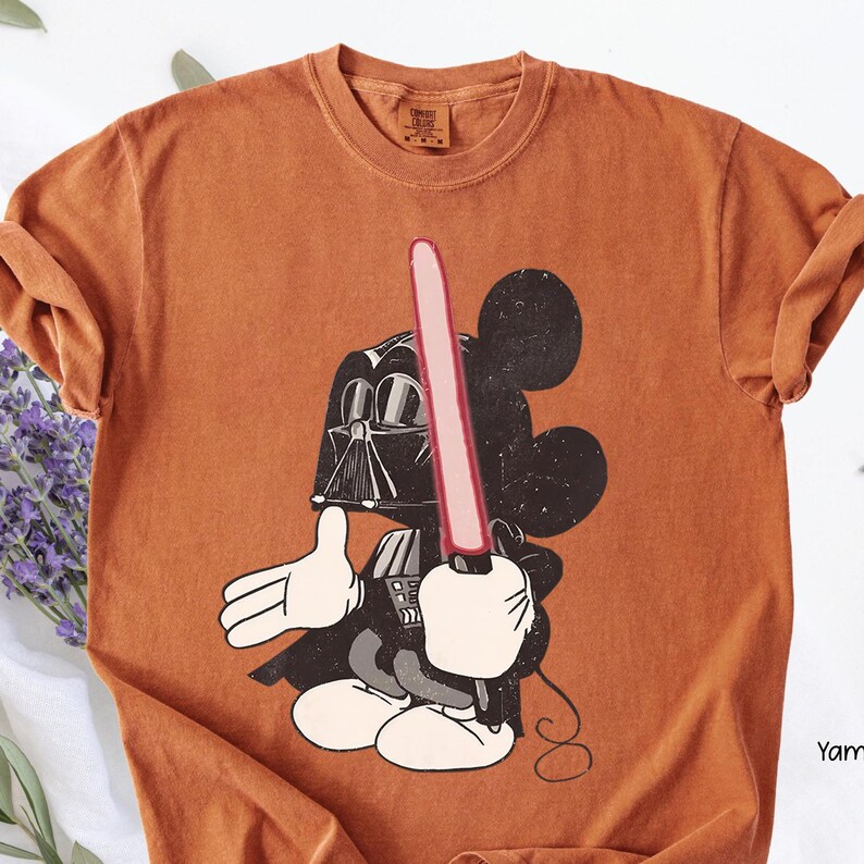 May include: Orange t-shirt featuring a graphic of Mickey Mouse dressed as Darth Vader, holding a red lightsaber. The design has a distressed, vintage look. The shirt is a Comfort Colors brand.
