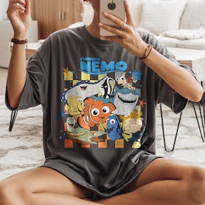 May include: Dark gray t-shirt featuring a colorful graphic of characters from the movie "Finding Nemo." The design includes Nemo, Dory, Crush, and other sea creatures. The words "Finding Nemo" are printed above the characters in blue.