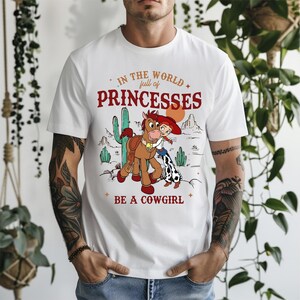 May include: White t-shirt with a graphic featuring a cartoon cowgirl and horse in a desert landscape. The text reads "IN THE WORLD full of PRINCESSES BE A COWGIRL" in red and orange lettering.