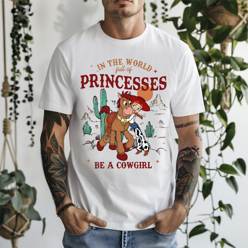 May include: White t-shirt with a graphic featuring a cartoon cowgirl and horse in a desert landscape. The text reads "IN THE WORLD full of PRINCESSES BE A COWGIRL" in red and orange lettering.