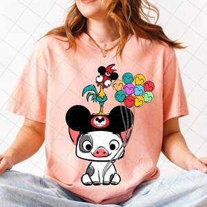 May include: A light pink t-shirt featuring a cartoon pig wearing a Mickey Mouse hat, with a rooster on top. The pig is holding a bunch of colorful balloons. The shirt has a crew neck and short sleeves.
