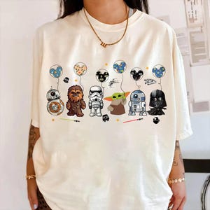 K&ouml;nnte beinhalten: Cremefarbenes T-Shirt mit Star Wars-Charakteren, die Mickey Mouse-f&ouml;rmige Ballons halten. Zu den Charakteren geh&ouml;ren BB-8, Chewbacca, ein Sturmtruppler, Grogu, R2-D2 und Darth Vader. Das Design umfasst Sterne und Lichtschwerter.
