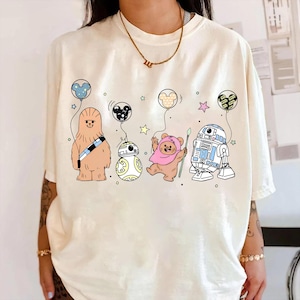 May include: Cream-colored t-shirt featuring cartoon characters from Star Wars and Mickey Mouse. The design includes Chewbacca, BB-8, an Ewok, and R2-D2, each holding a balloon. The balloons are in the shape of Mickey Mouse heads and stars.