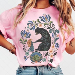 May include: Pink t-shirt featuring a black bear holding hands with a girl, surrounded by a floral wreath. The design includes blue, green, and yellow floral elements. The shirt is a crew neck style.