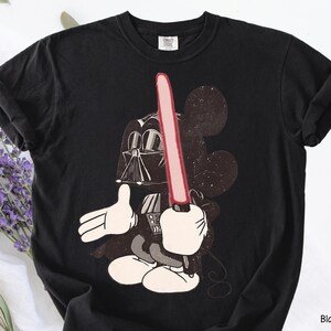 May include: Black t-shirt featuring a graphic of Mickey Mouse as Darth Vader, holding a red lightsaber. The design includes Mickey's iconic ears and white gloves. The shirt is made of a soft material.