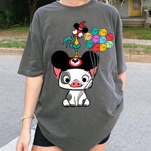 May include: Dark gray t-shirt featuring a cartoon pig character with Mickey Mouse ears, a rooster, and colorful balloons. The pig is white with black spots and a pink snout. The rooster is yellow, red, and blue.