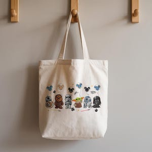 K&ouml;nnte beinhalten: Cremefarbene Tragetasche mit Cartoon-Star-Wars-Figuren, die Mickey-Mouse-f&ouml;rmige Luftballons halten. Zu den Figuren geh&ouml;ren BB-8, Chewbacca, ein Sturmtruppler, Yoda, R2-D2 und Darth Vader. Die Tasche h&auml;ngt an Holzhaken an einer grauen Wand.