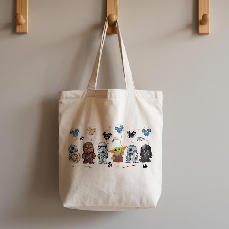 K&ouml;nnte beinhalten: Cremefarbene Tragetasche mit Cartoon-Star-Wars-Figuren, die Mickey-Mouse-f&ouml;rmige Luftballons halten. Zu den Figuren geh&ouml;ren BB-8, Chewbacca, ein Sturmtruppler, Yoda, R2-D2 und Darth Vader. Die Tasche h&auml;ngt an Holzhaken an einer grauen Wand.