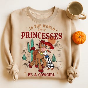 May include: A tan sweatshirt featuring a graphic of a cartoon cowgirl hugging a horse, with a desert landscape backdrop. The text reads "In the world full of princesses, be a cowgirl." A mug and small pumpkin are on the right.