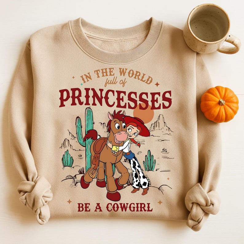May include: A tan sweatshirt featuring a graphic of a cartoon cowgirl hugging a horse, with a desert landscape backdrop. The text reads "In the world full of princesses, be a cowgirl." A mug and small pumpkin are on the right.