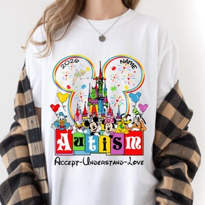 May include: White t-shirt featuring a colorful Disney-themed design. The design includes a castle, fireworks, and various Disney characters. The word "Autism" is prominently displayed, along with the phrase "Accept-Understand-Love". The year "2026" is also visible.