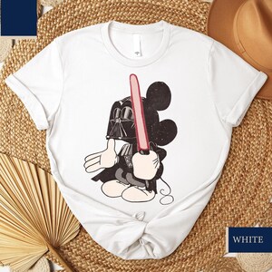 May include: White t-shirt featuring a cartoon Mickey Mouse character dressed as Darth Vader, holding a red lightsaber. The design is in black and white with red accents. The shirt is a crew neck.