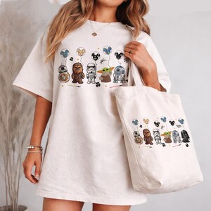 K&ouml;nnte beinhalten: Cremefarbenes T-Shirt und Tragetasche mit einem Star Wars-Design. Das Shirt und die Tragetasche zeigen Zeichentrickfiguren wie BB-8, Chewbacca, einen Sturmtruppler, Grogu und R2-D2, die jeweils einen Ballon halten. Das Design enth&auml;lt kleine Fahrzeuge.