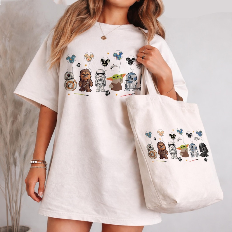 K&ouml;nnte beinhalten: Cremefarbenes T-Shirt und Tragetasche mit einem Star Wars-Design. Das Shirt und die Tragetasche zeigen Zeichentrickfiguren wie BB-8, Chewbacca, einen Sturmtruppler, Grogu und R2-D2, die jeweils einen Ballon halten. Das Design enth&auml;lt kleine Fahrzeuge.