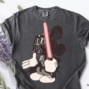 May include: Dark gray t-shirt featuring a cartoon Mickey Mouse character dressed as Darth Vader, holding a red lightsaber. The graphic is a mashup of the iconic Disney and Star Wars franchises. The shirt has a crew neck and short sleeves.