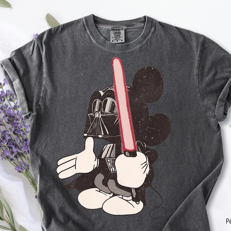 May include: Dark gray t-shirt featuring a cartoon Mickey Mouse character dressed as Darth Vader, holding a red lightsaber. The graphic is a mashup of the iconic Disney and Star Wars franchises. The shirt has a crew neck and short sleeves.